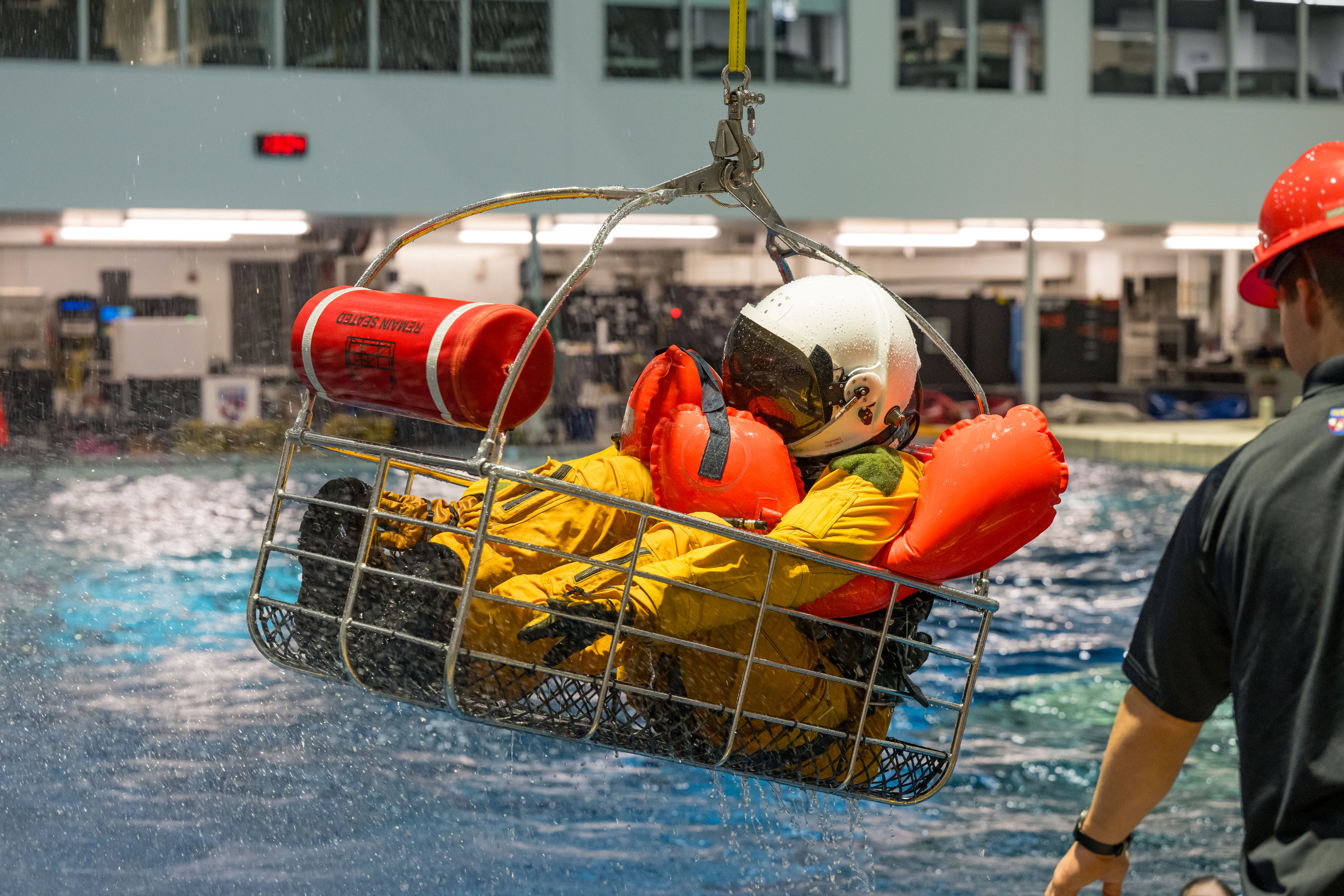 A person in a yellow flight suit and white helmet sits in an airlift rescue basket, hovering above the Neutral Buoyancy Laboratory pool.