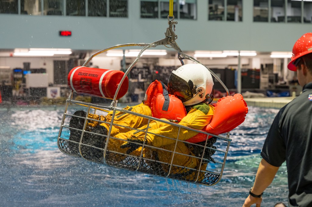 A person in a yellow flight suit and white helmet sits in an airlift rescue basket, hovering above the Neutral Buoyancy Laboratory pool.