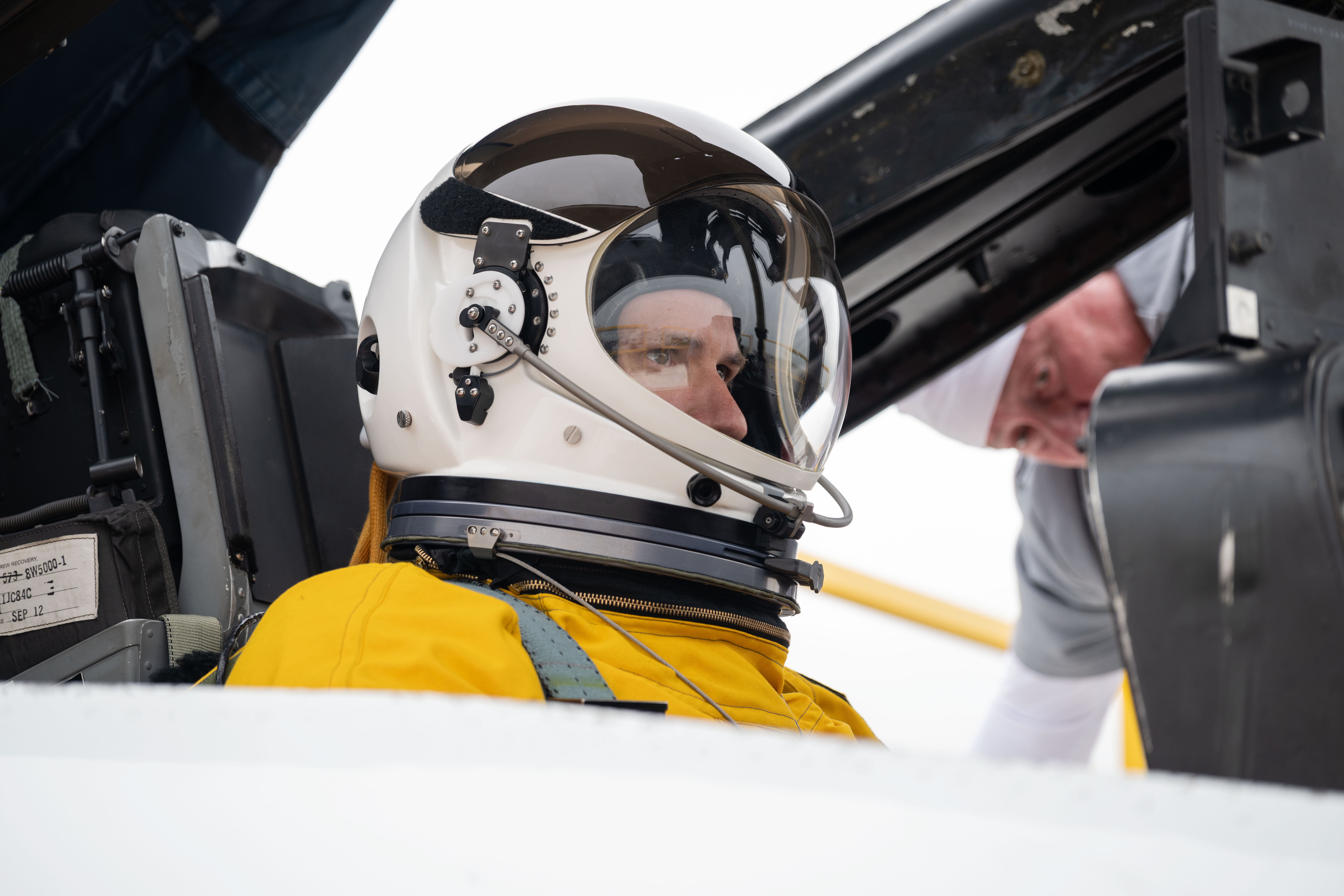 A man wearing a yellow flight suit and white helmet is shown, seated inside the cockpit of a WB-57 aircraft.