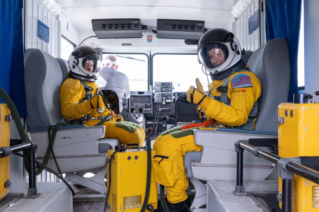 Two people wearing yellow flight suits and white helmets ride in the back of a van.