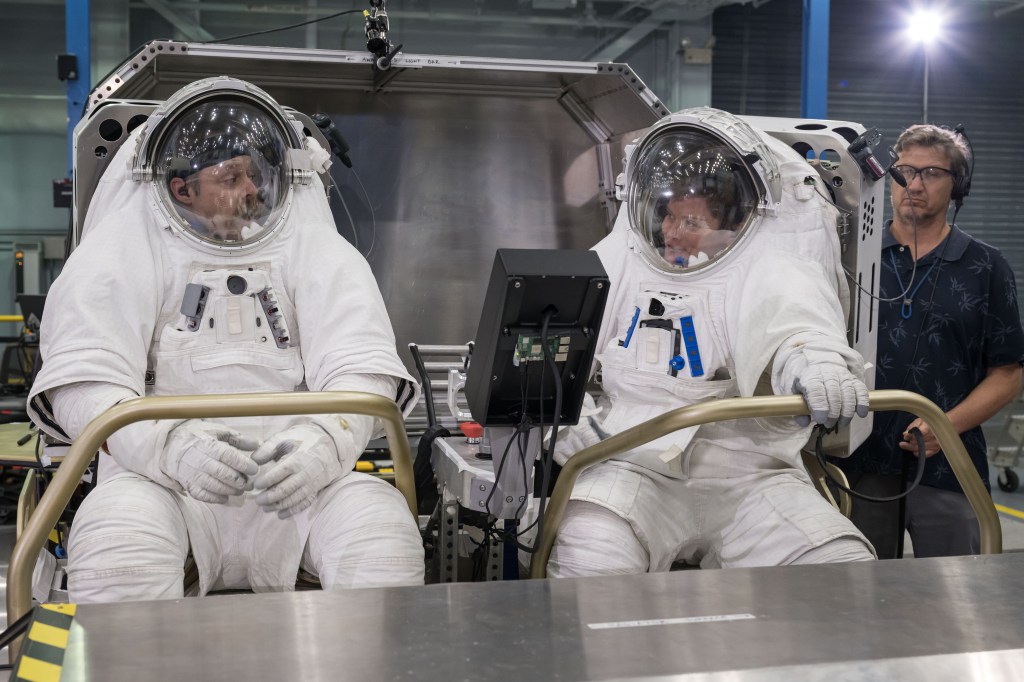NASA astronaut Anne McClain, right, and a NASA test engineer sit side by side in white spacesuits aboard Lunar Outpost’s Pegasus Lunar Terrain Vehicle prototype during human-in-the-loop testing. A display and controls are positioned between them. A man wearing a headset stands behind the vehicle. Credit: NASA