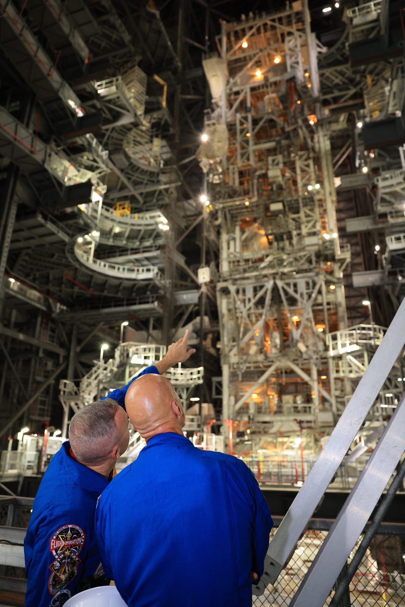 Two people in blue flight suits look upward at a tall, complex structure made of pipes, scaffolding, and machinery.