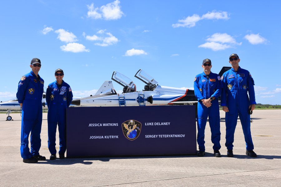 Four astronauts in blue flight suits stand next to a banner in front of NASA jets on a runway.