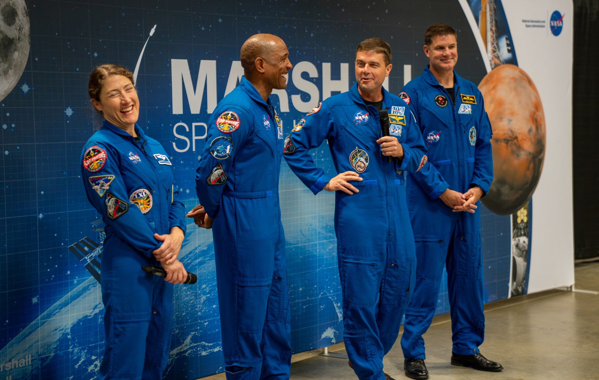 The Artemis II crew smile and laugh. They are wearing blue jumpsuits with patches on them. Christina Koch, left, and Reid Wiseman, second from right, are holding microphones.
