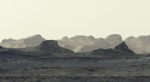 A stark, grayish-tan Martian landscape features jagged rock formations and rolling mountain ridges beneath a pale, hazy sky.