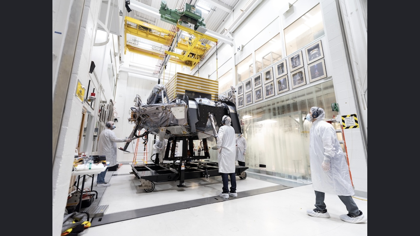 Engineers in white clean-room garb monitor a large spacecraft as it is hoisted by a yellow overhead crane in a hallway in a bright, high-tech laboratory. Framed photos line both sides of the hallway.