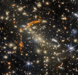 A massive galaxy cluster, made of many golden elliptical galaxies with the largest, brightest one in the center. Heavily distorted orange galaxies appear all around the cluster, the largest concentrated in a large arc around the cluster's center; these are images of very distant galaxies, created by gravitational lensing. A few galaxies are large and bluish-white in color; bright, nearby stars are the same color.