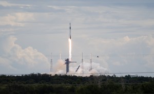 A SpaceX Falcon 9 rocket carrying the company's Dragon spacecraft launches. There is a bright column of fire under the rocket as it moves upward. Clouds of vapor spread out below the rocket, almost blending into the similarly shaped clouds in the sky.