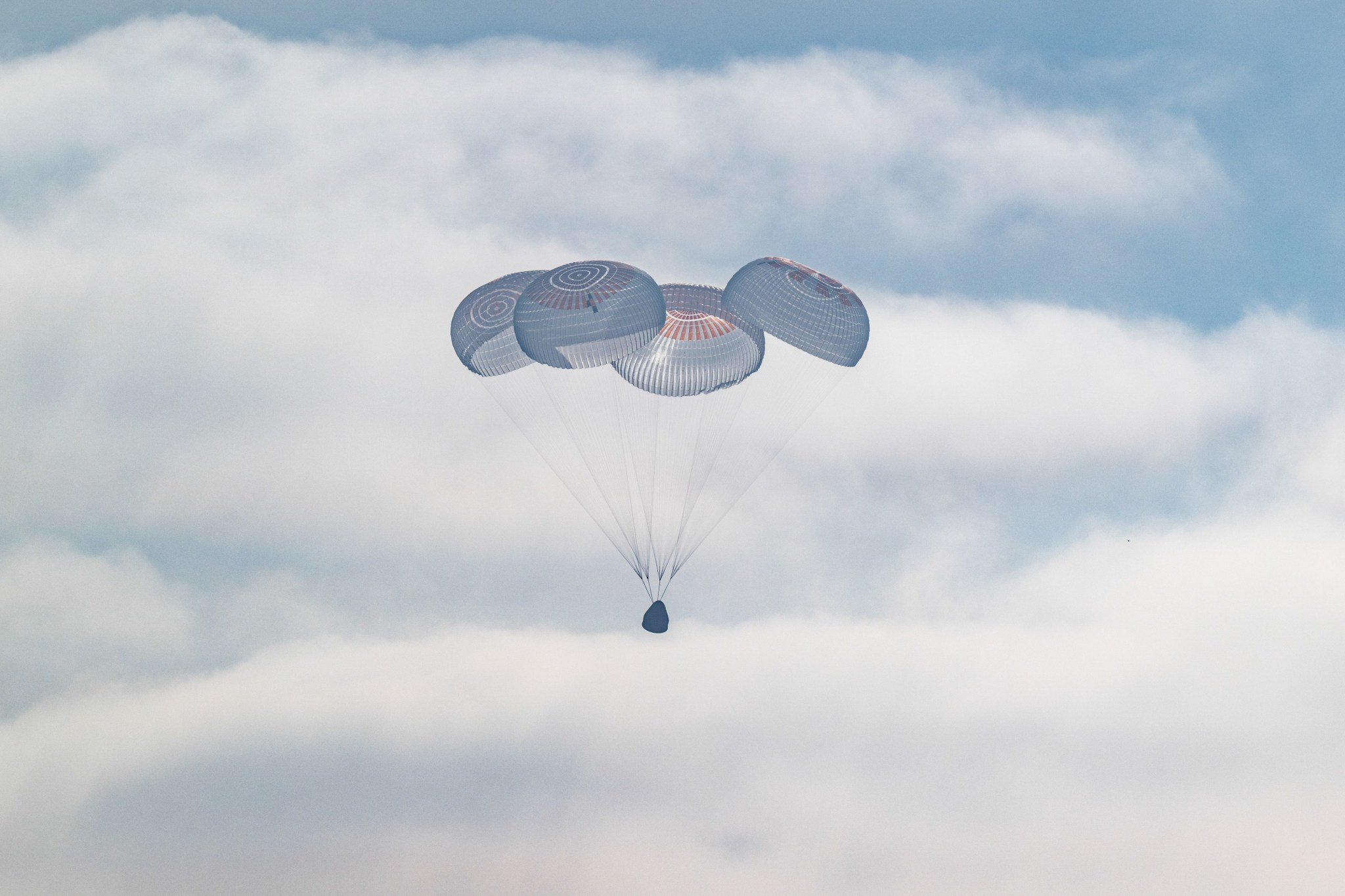A cone-shaped spacecraft and the four large parachutes attached to it against a backdrop of fluffy white clouds in the sky.