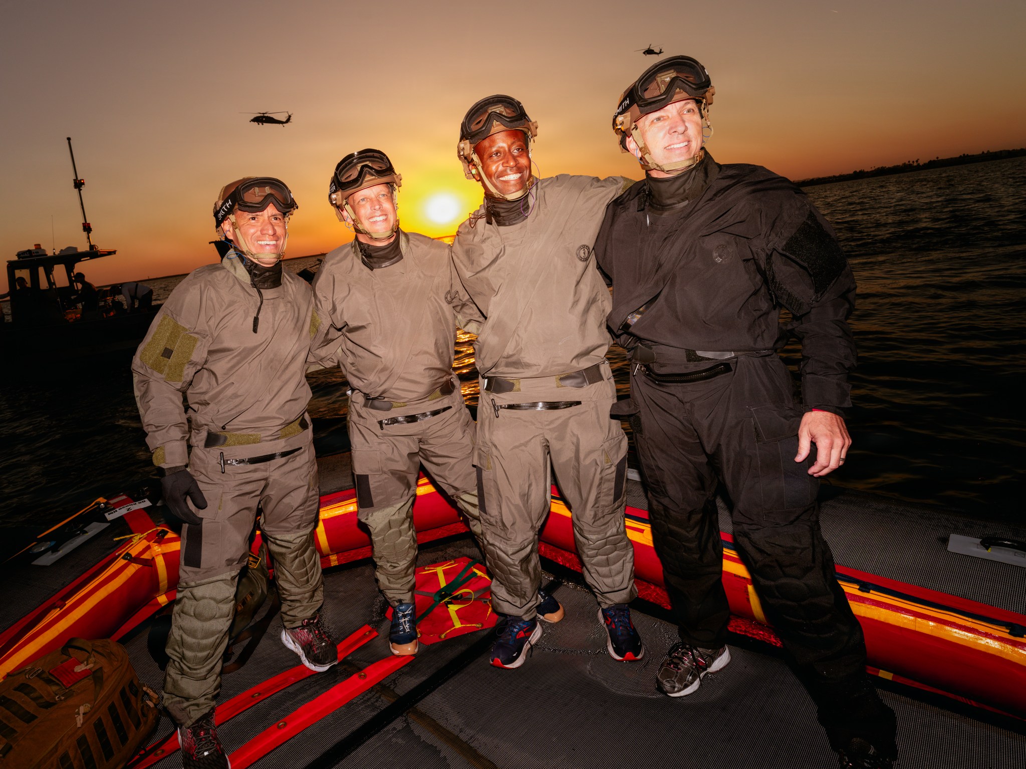 Image shows four men in brown water training suits standing near water. Behind them is a US Navy boat and helicopter with a sunset.