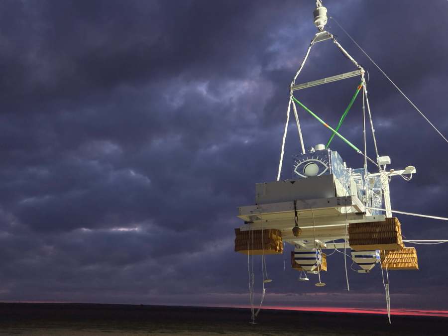 A balloon gondola suspended in the air against a dark cloud sky.