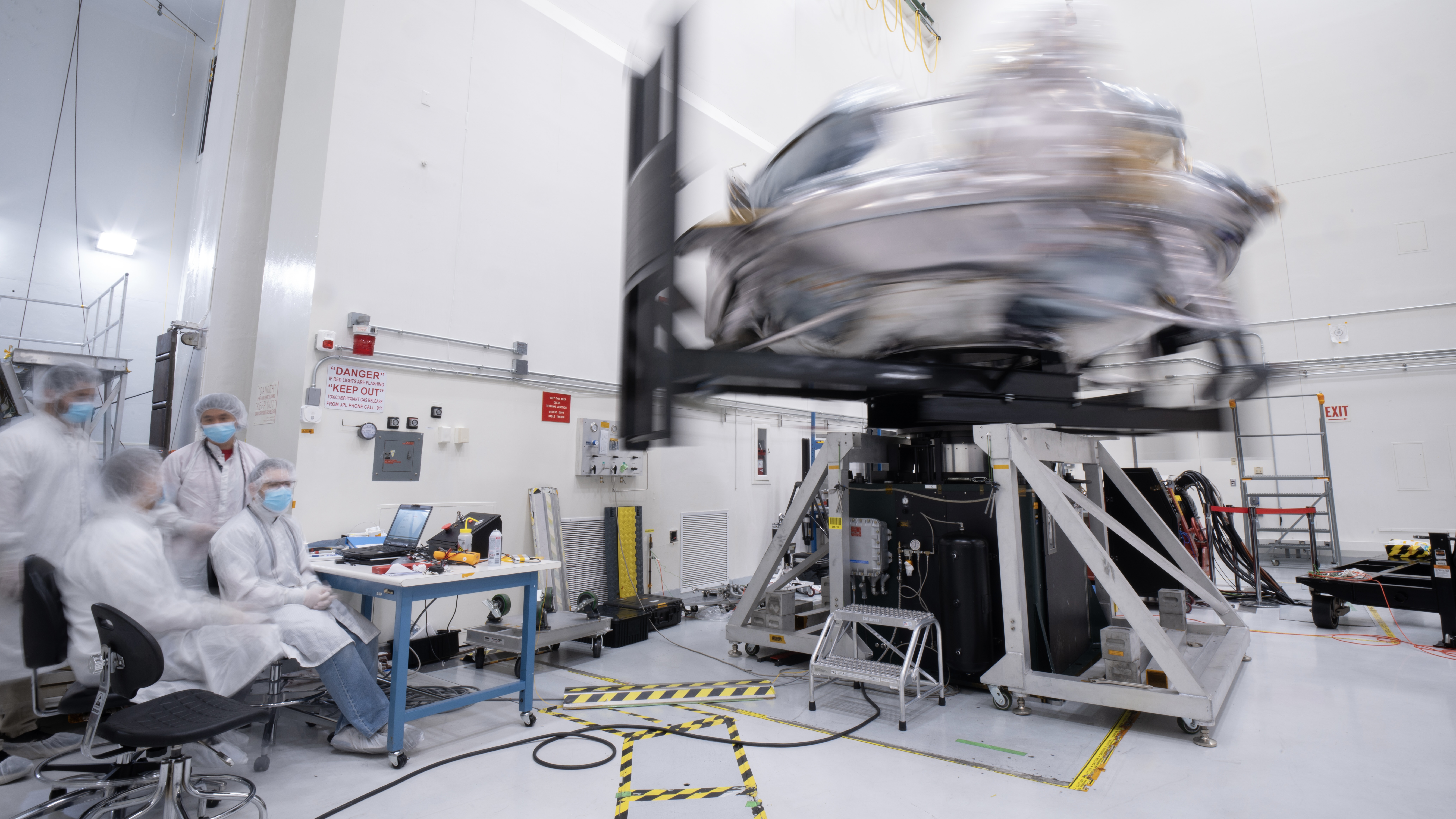 Engineers in white clean-room frocks monitor a silvery spacecraft spinning so rapidly it’s blurred on a test rig inside a bright laboratory.