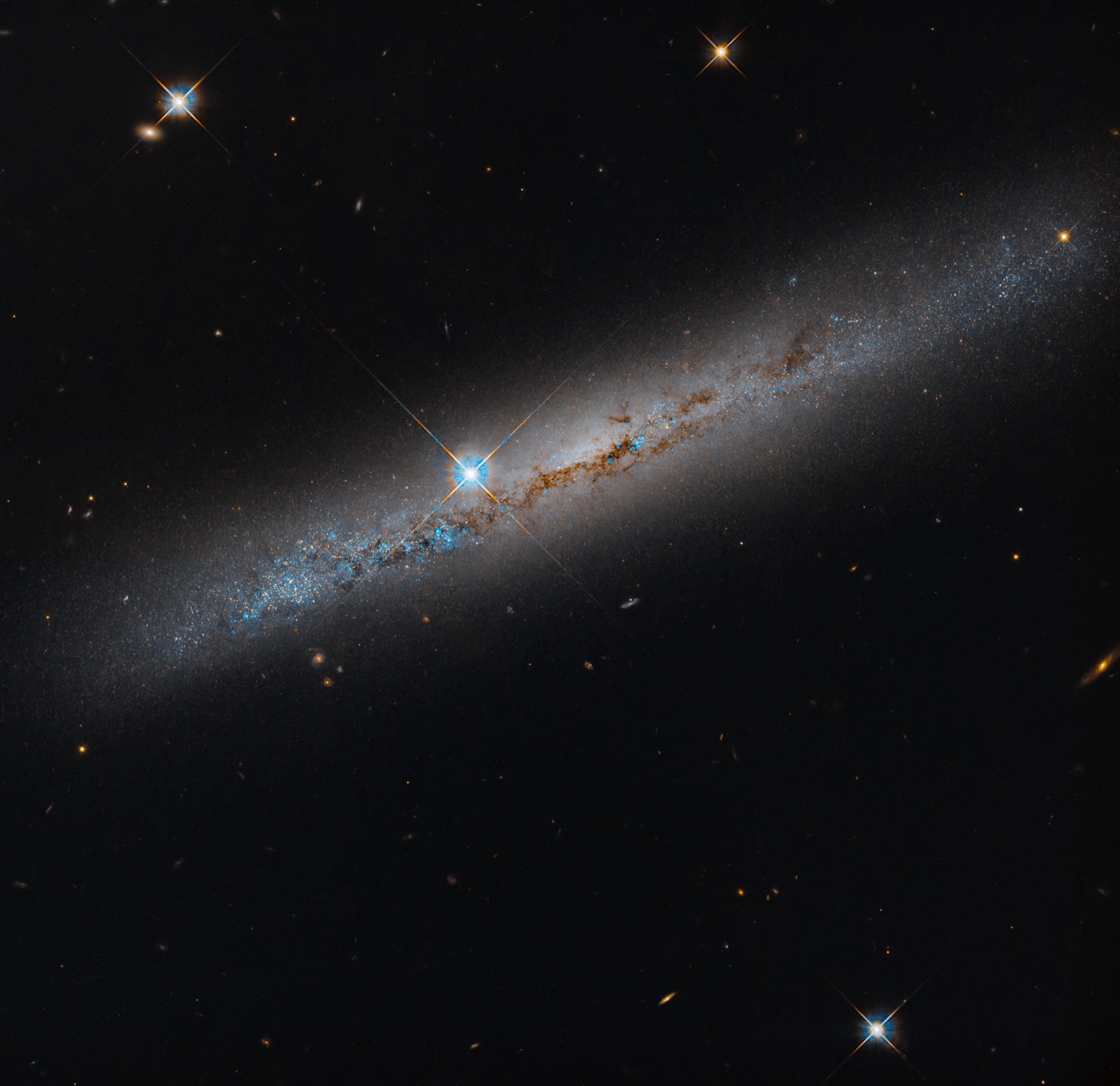 An edge-on spiral galaxy. Its rusty-brown dust lanes bisect the galaxy lengthwise. Bright-blue points dot the galaxy and seem to be more dense on its left side. A bright foreground star shines just to the left of the galaxy's center. The background is black and dotted with distant galaxies and a smattering of foreground stars, which are easily identified by their diffraction spikes.