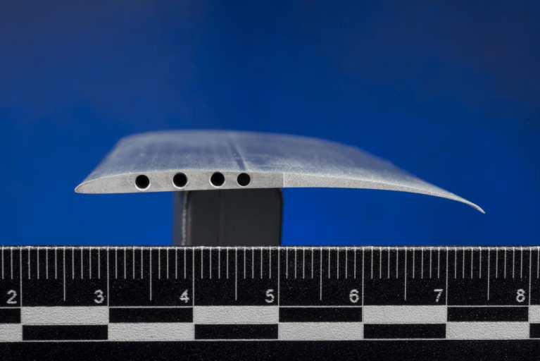 A close-up view of a cross-section of the Ingenuity rotor blade, also called an airfoil. The blade is shaped like an arched teardrop with holes drilled into the side.