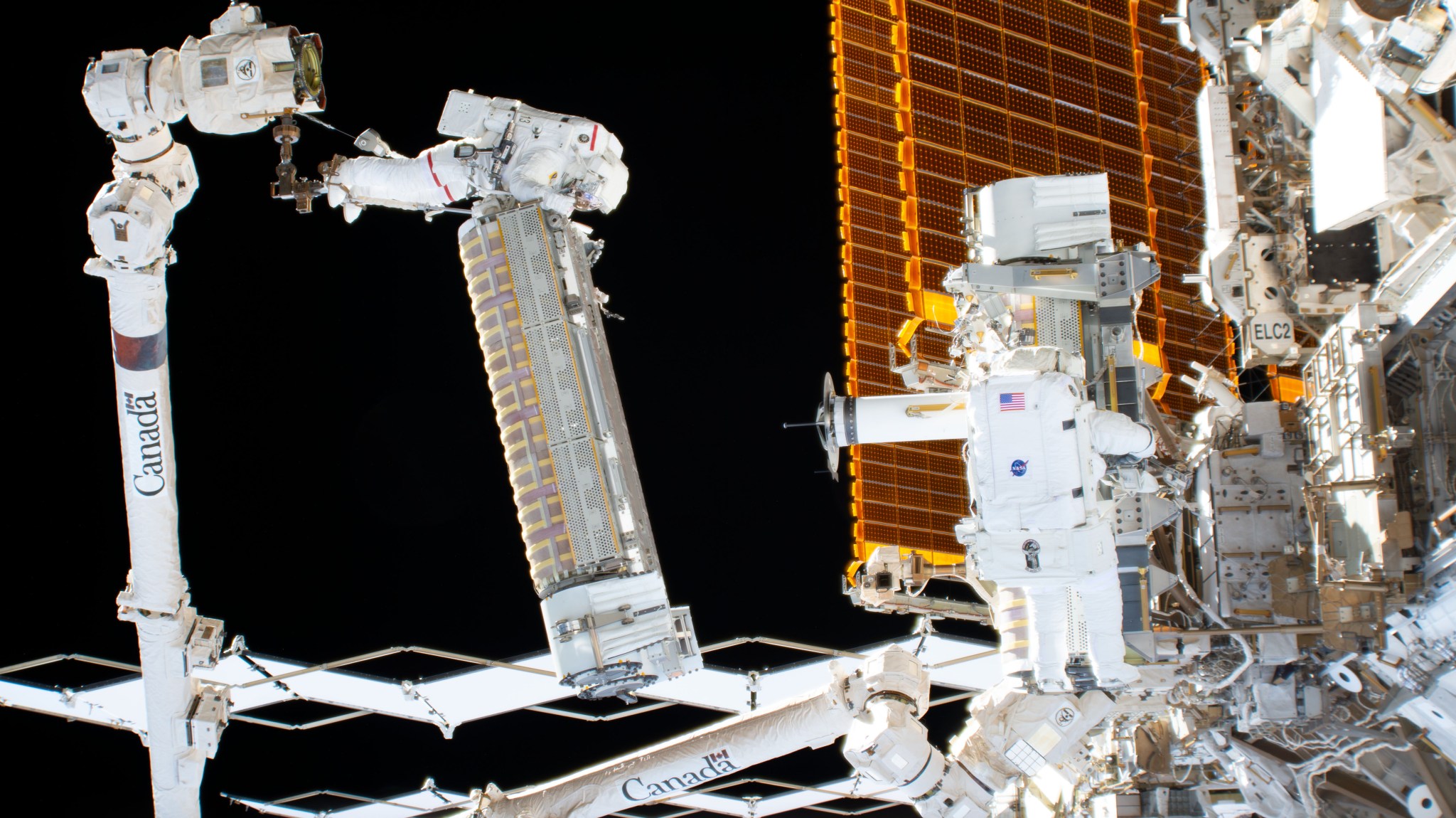 Two NASA astronauts in white spacesuits work outside the International Space Station, with one holding a rectangular hardware unit while robotic arms assist near the station’s orange solar arrays.