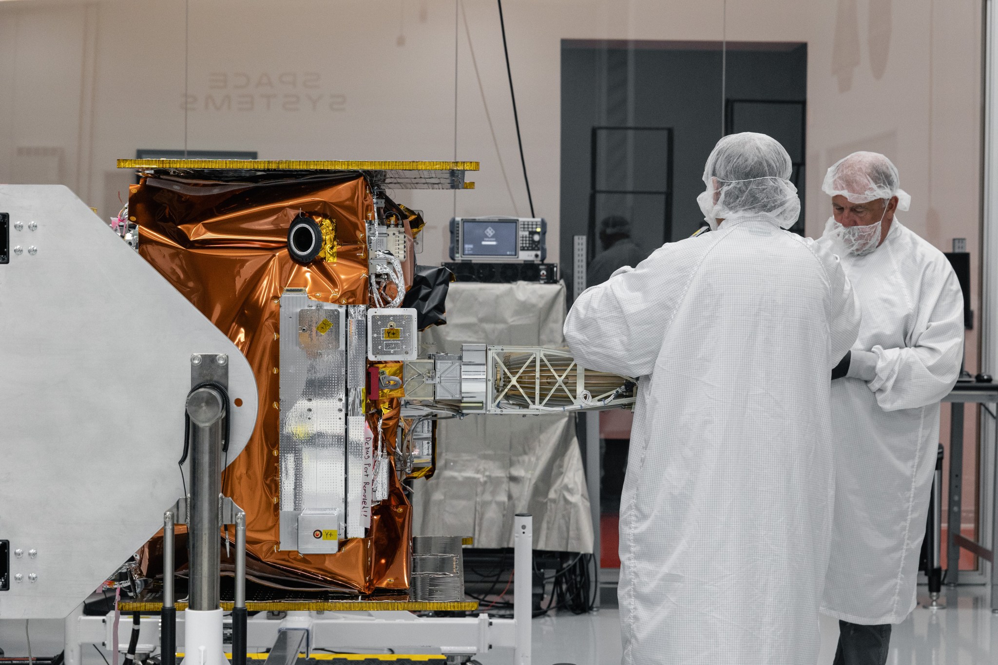 Technicians wearing cleanroom garments and hair coverings work on a satellite inside a spacecraft processing facility. The satellite, wrapped in gold-colored thermal insulation and mounted on support equipment, is positioned in the foreground while personnel inspect and handle an extended structural component.