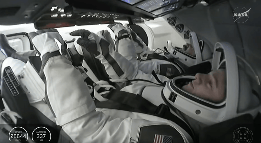 Image is inside a spacecraft with astronauts wearing white and black spacesuits.