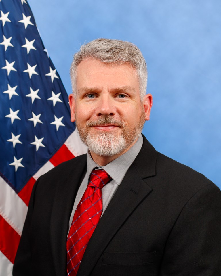 Portrait of Shad Combs standing in front of an American flag and back ground. He is wearing a black blazer jacket, red tie, and grey undershirt.