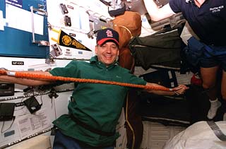 Mission specialist Tom Akers, holding a bunge cord in both hands and wearing a baseball cap, stretches his arms out the width of the Atlantis's middeck.