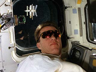 STS-81 pilot Brent Jett (wearing sunglasses) is photographed on the Atlantis's aft flight deck during the approach to the Mir space station. The Mir is visible in the overhead window.