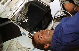 Mission specialist Jean-Francois Clervoy with laser targeting equipment in shuttle's flight deck with a distant Mir in the overhead window.