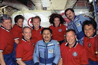 STS-91 and Mir-25 crewmembers pose for photos in the Mir Base Block module with peace banners behind them.